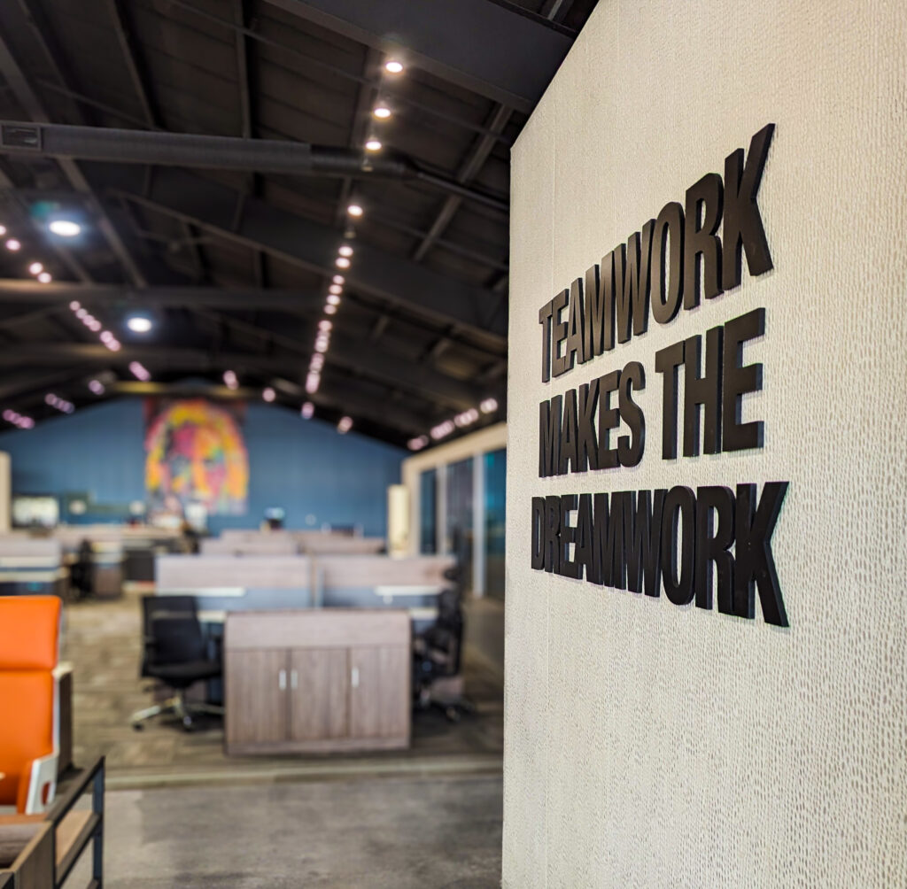 A photo of a wall with the words "Teamwork makes the dreamwork" on it, with Oakbay in the background